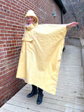 Woman standing a yellow rain poncho by a brick wall with her arm to the side to show how wide the poncho is.